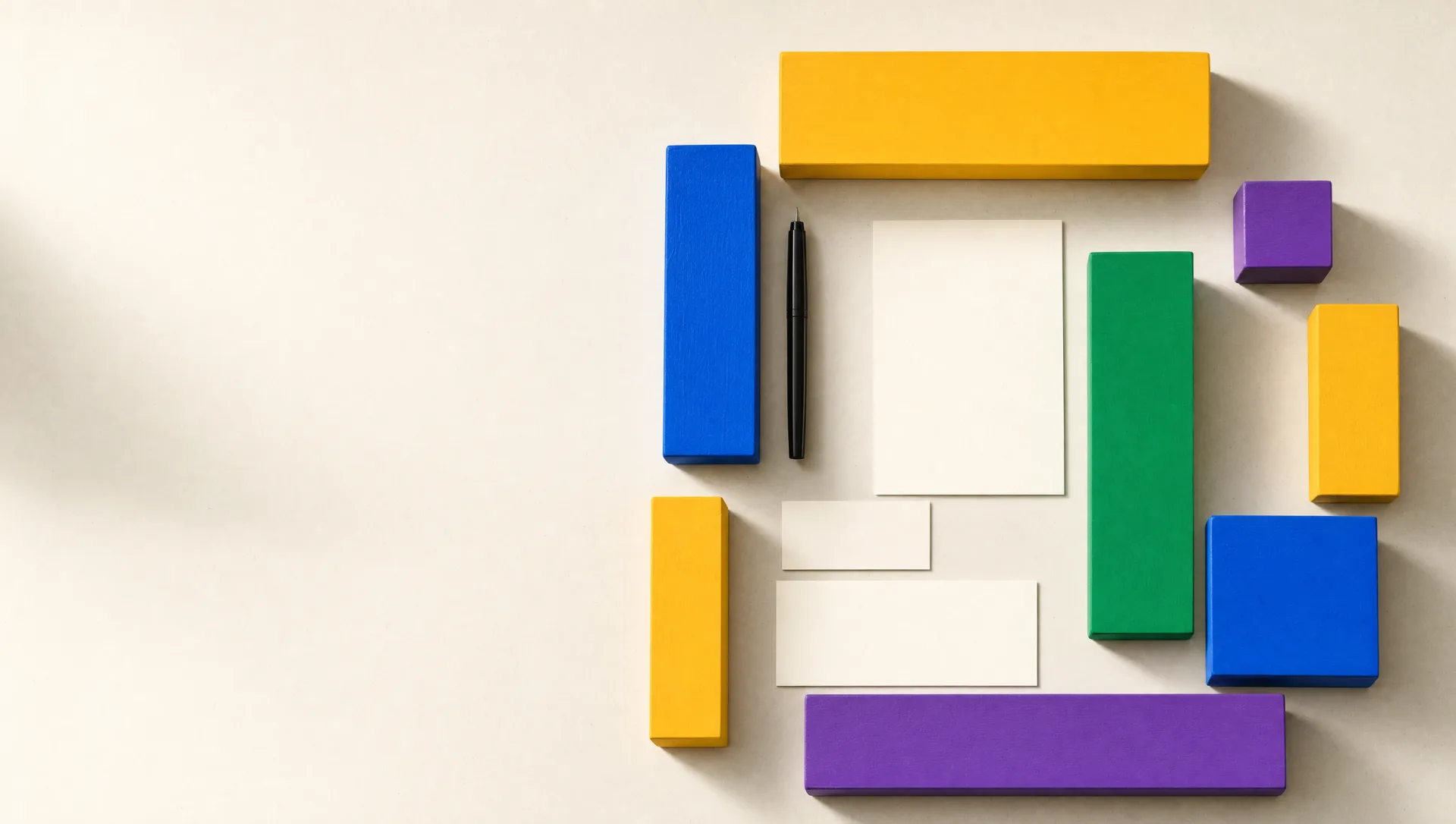 Painted wooden blocks in yellow, blue, green and violet arranged in a neat grid on warm off-white paper, with blank cream note cards and a black pen among them.